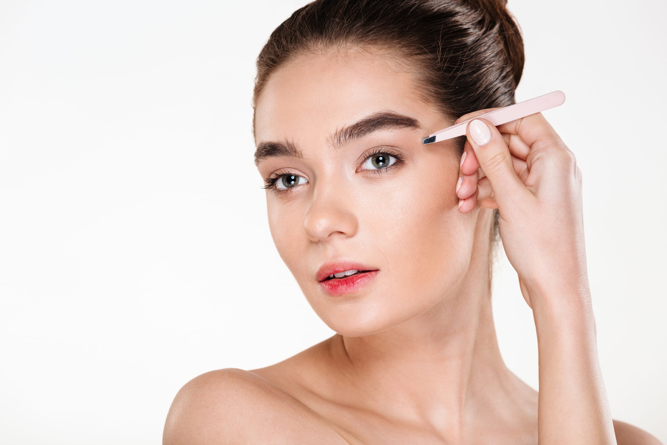 Close up studio portrait of young brunette woman with soft skin plucking eyebrows with tweezers, isolated over white background
